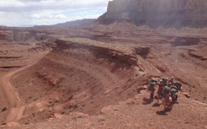 A group of students wearing backpacks huddle together on a mesa overlooking a red desert landscape